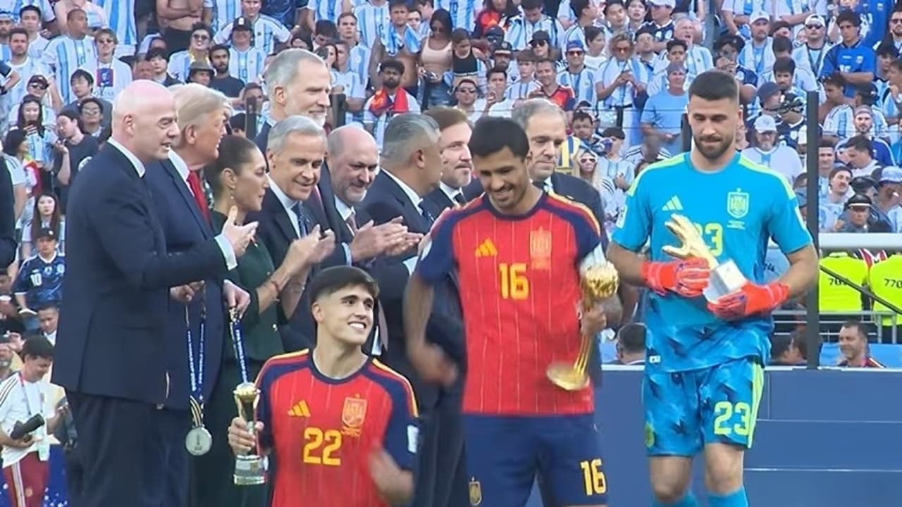 Sur le podium du MetLife Stadium, Rodri, maillot rouge numéro 16, tient le trophée du Ballon d'or à côté de Pau Cubarsi, numéro 22, et du gardien Unai Simon en tenue bleue, devant Gianni Infantino et les officiels qui applaudissent.