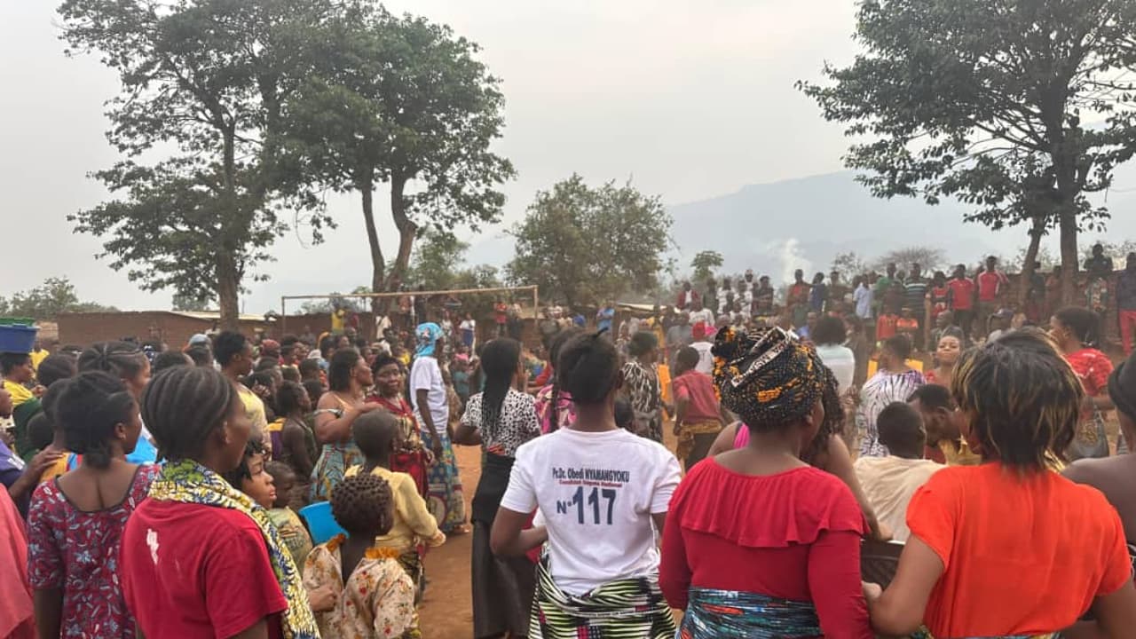 Rassemblement de villageois, surtout des femmes en pagnes colorés, sur une place de terre battue d'un village des hauts plateaux de Fizi, montagnes en arrière-plan ; au premier plan, une femme porte un t-shirt à l'effigie du député Obedi Nyamangyoku.