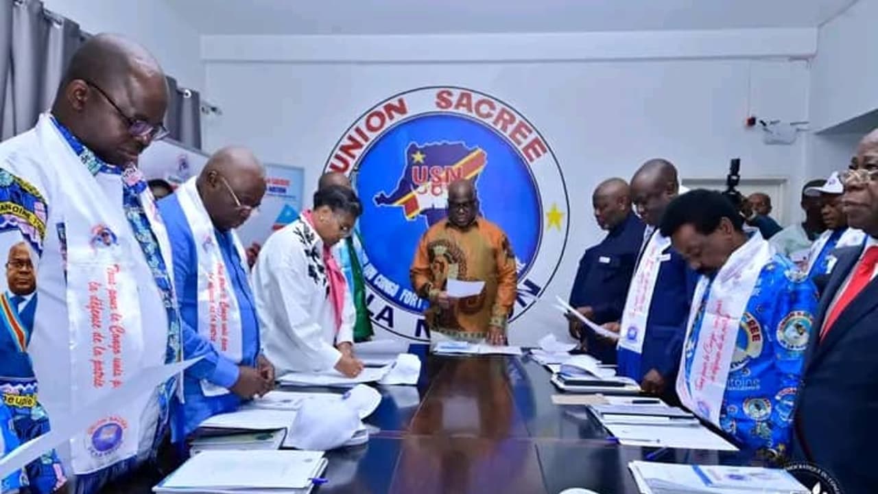Des cadres de l'Union sacrée de la nation, écharpes aux couleurs de la plateforme, debout autour d'une table de réunion devant le logo de l'USN, à Kinshasa