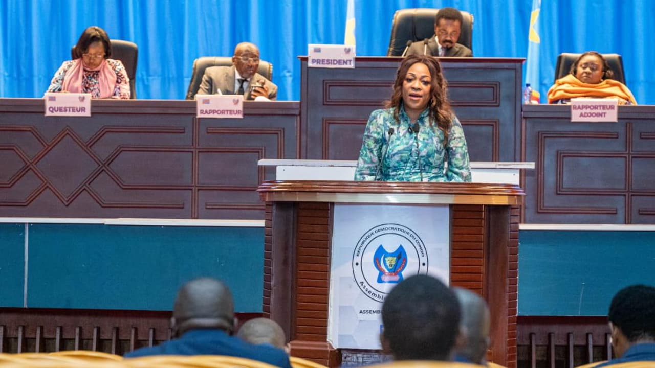 La députée Dorothée Madiya Mwamba, en veste à motifs verts, s'exprime à la tribune de l'Assemblée nationale de la RDC, derrière un pupitre portant l'emblème de l'institution ; les membres du présidium sont assis en surplomb.