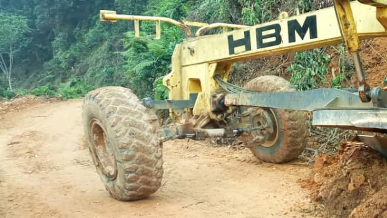 Une niveleuse jaune de marque HBM, aux gros pneus couverts de boue, travaille sur une route de terre fraîchement ouverte, taillée à flanc de colline à travers la forêt dense ; la terre ocre retournée s'entasse sur le bas-côté.