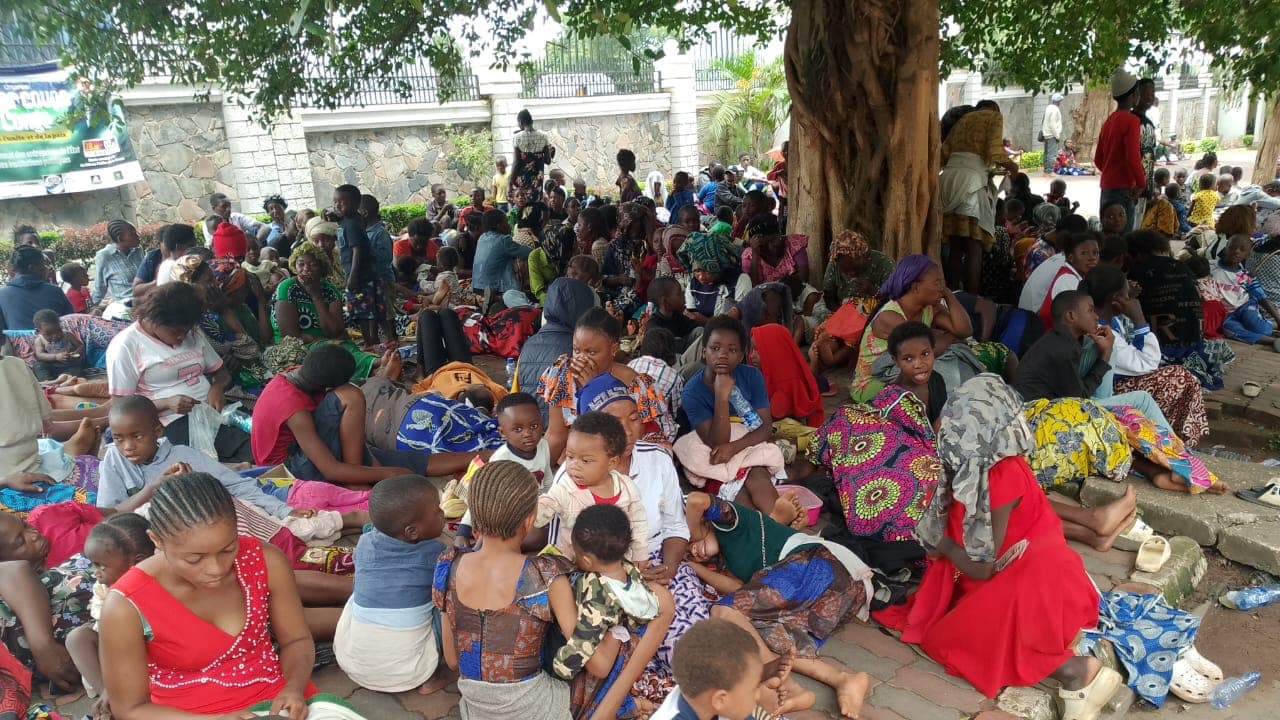 Des familles déplacées de l'Est, femmes et enfants, assises à même le sol devant le mur du gouvernorat du Haut-Katanga à Lubumbashi, une banderole de leur collectif visible en arrière-plan.