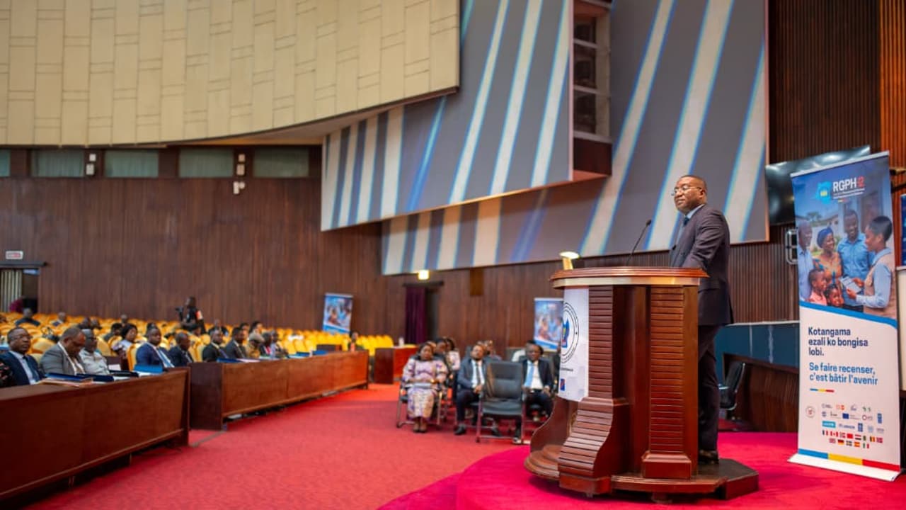 Guylain Nyembo s'exprime depuis un pupitre dans l'hémicycle du Palais du Peuple à Kinshasa, devant une assistance assise, à côté d'un kakémono du RGPH-2 portant le slogan « Se faire recenser, c'est bâtir l'avenir »