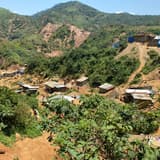 Vue en plongée du site minier artisanal de Lupupu, à Kamituga : des cabanes en planches et en tôle dispersées sur des collines éventrées par des excavations de terre ocre, entourées de végétation verte, avec des creuseurs au travail.