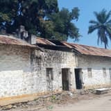 Bâtiment bas de la prison centrale de Feshi, murs de maçonnerie blanchis et fissurés, toiture en tôles rouillées partiellement arrachée, palmiers en arrière-plan