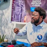 Clovis Mutsuva, en chemise bleu clair portant un badge du parti Ensemble pour la République, s'exprime devant plusieurs micros de presse, devant une banderole à son nom.