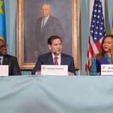 Trois responsables assis derrière une table recouverte d'une nappe bleue, devant des drapeaux du Rwanda, des États-Unis et de la République démocratique du Congo. Des plaques nominatives posées devant eux indiquent « Rwanda », « United States » et « Democratic Republic of the Congo ». À gauche, un homme en costume sombre portant des lunettes ; au centre, un homme en costume à cravate rouge qui parle dans un micro ; à droite, une femme en veste bleue et tenue en pagne coloré. Un portrait peint est accroché au mur derrière eux.