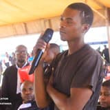 Un jeune homme debout, un micro à la main, répond aux questions du tribunal militaire de garnison de Lubumbashi lors d'une audience foraine sous une tente ; des avocats en robe noire et un militaire en béret bleu l'entourent.