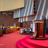 Guylain Nyembo s'exprime depuis un pupitre dans l'hémicycle du Palais du Peuple à Kinshasa, devant une assistance assise, à côté d'un kakémono du RGPH-2 portant le slogan « Se faire recenser, c'est bâtir l'avenir »