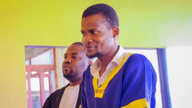 Nathanaël Onokomba, vêtu de la tenue bleu et jaune des détenus, se tient debout aux côtés d'un avocat en robe noire dans une salle aux murs verts.