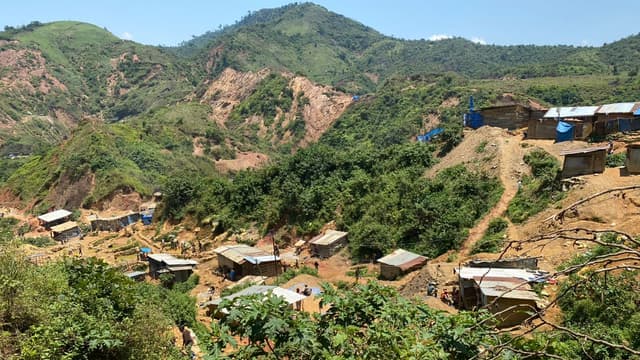 Vue en plongée du site minier artisanal de Lupupu, à Kamituga : des cabanes en planches et en tôle dispersées sur des collines éventrées par des excavations de terre ocre, entourées de végétation verte, avec des creuseurs au travail.