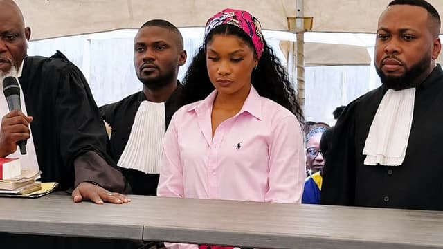 La chanteuse Rebo Tchulo, chemise rose et foulard fleuri, se tient debout derrière une table entre ses avocats en robe noire lors d'une audience du tribunal militaire.