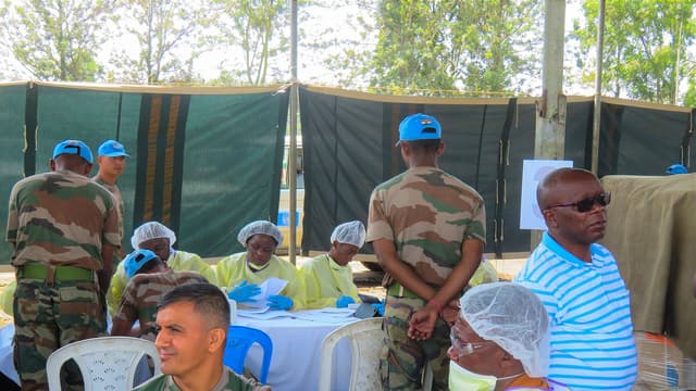 Séance de vaccination contre Ebola en plein air en République démocratique du Congo : des agents de santé congolais en blouse jaune, gants bleus, masque et charlotte administrent un vaccin dans le bras d'un homme assis, tandis que d'autres soignants enregistrent les personnes vaccinées à une table.
