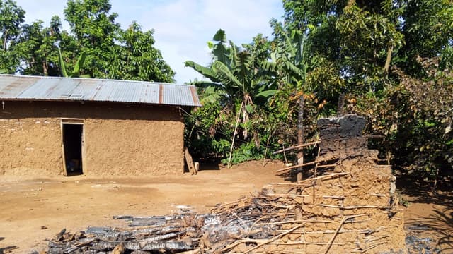 Une maison en briques de terre au toit de tôle et, au premier plan, les décombres calcinés d'une habitation incendiée, dans un village rural entouré de bananiers et de forêt, après une attaque des ADF.