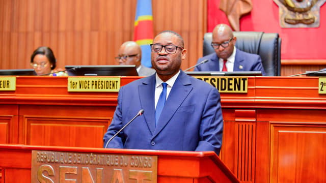 Le ministre congolais des Finances Doudou Fwamba, en costume bleu, s'exprime debout au pupitre marqué « République démocratique du Congo , Sénat », devant les membres du bureau assis à la tribune.