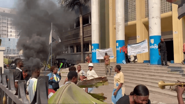 Agents de la SCPT en grève devant le siège de l'entreprise à Kinshasa : fumée noire de pneus brûlés, banderoles réclamant le départ de la direction, manifestants avec vuvuzelas et trompette.
