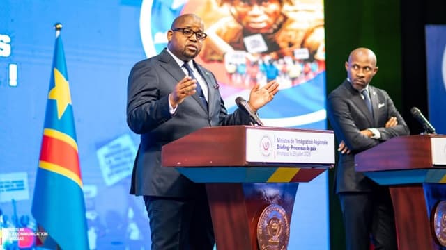Floribert Anzuluni, ministre de l'Intégration régionale, s'exprime les mains ouvertes derrière un pupitre marqué « Ministre de l'Intégration Régionale, Briefing - Processus de paix, Kinshasa, le 28 juillet 2026 » ; à droite, Patrick Muyaya, bras croisés, à son propre pupitre, devant le drapeau de la RDC.