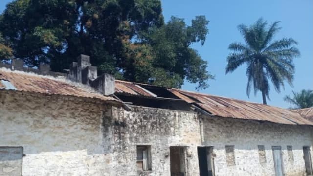 Bâtiment bas de la prison centrale de Feshi, murs de maçonnerie blanchis et fissurés, toiture en tôles rouillées partiellement arrachée, palmiers en arrière-plan