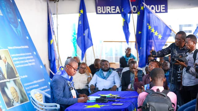 Un responsable du PALU lit une déclaration derrière une table drapée du drapeau bleu à étoiles jaunes du parti, entouré de drapeaux du PALU, de militants assis et de journalistes, sous une banderole « Bureau politique », au siège du parti à Kinshasa.