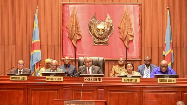 Le présidium du Sénat congolais au Palais du Peuple : Jean-Michel Sama Lukonde, en costume bleu et lunettes, siège au centre sous le pupitre marqué PRÉSIDENT, entouré des vice-présidents, rapporteurs et questeur, devant l'emblème du Sénat et deux drapeaux de la RDC.