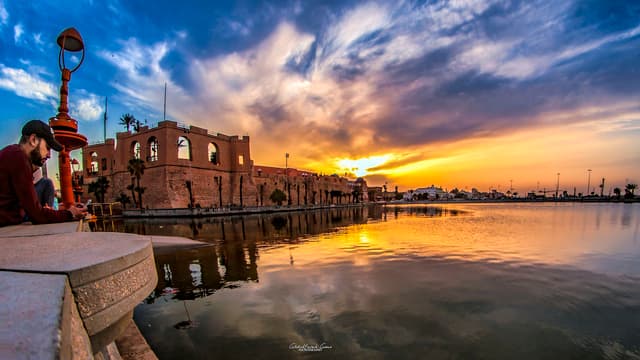 Le château rouge de Tripoli, forteresse de pierre ocre illuminée au coucher du soleil, bordant un plan d'eau qui reflète le ciel orangé ; la ville s'étend à l'arrière-plan.