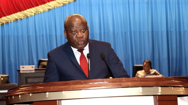 Un homme en costume bleu marine et cravate rouge parle dans deux micros, debout à la tribune en bois de l'Assemblée nationale congolaise, devant une tenture bleue et le drapeau de la RDC.