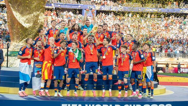 Les joueurs espagnols réunis sur le podium soulèvent le trophée de la Coupe du monde devant un panneau « FIFA World Cup 2026 Champions », sous les confettis.
