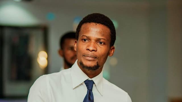 Portrait de Nathanaël Onokomba, chemise blanche et cravate bleu marine ornée d'une épinglette en forme de carte de la République démocratique du Congo