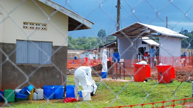 Agents de santé ougandais en combinaison de protection intégrale blanche au centre de traitement Ebola de l'hôpital régional de Mubende : l'un désinfecte du matériel au-dessus de seaux, tentes d'isolement bâchées et poubelles rouges à déchets biologiques à l'arrière-plan, derrière une clôture grillagée et un filet de sécurité orange.