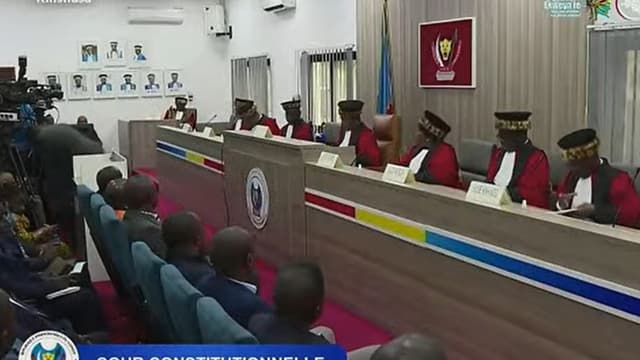 Les juges de la Cour constitutionnelle de la RDC, en toge rouge et toque bordée de peau de léopard, siègent en audience publique à Kinshasa, devant les armoiries de la République ; au premier plan, l'assistance et les caméras.