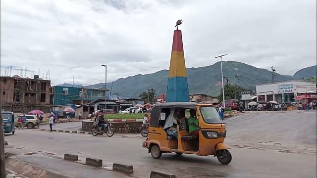 Scène de rue au centre-ville d'Uvira : un bajaj jaune et des motos passent devant un rond-point orné d'un monument aux couleurs du drapeau congolais, bordé de boutiques, avec les montagnes en arrière-plan.