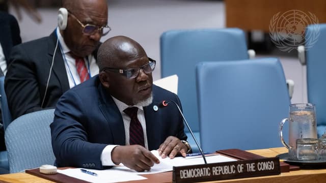 Un homme en costume sombre et lunettes, assis au Conseil de sécurité de l'ONU derrière une plaque « Democratic Republic of the Congo », s'exprime en tenant des documents, sous le logo des Nations unies.