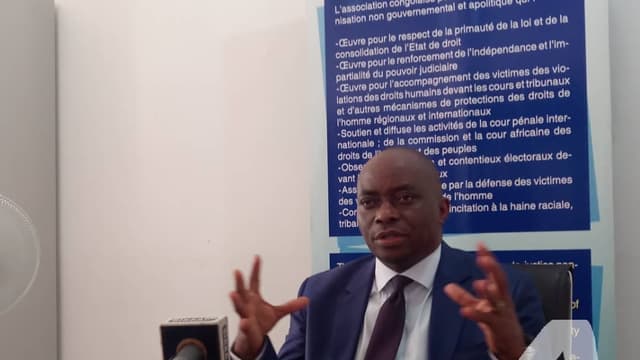 Georges Kapiamba, président de l'ACAJ, en costume sombre, assis derrière une table garnie de microphones, gesticulant des deux mains lors d'une conférence de presse, devant une banderole bleue de l'association.