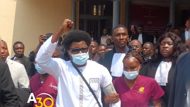 Constant Mutamba, le poing levé, portant un masque chirurgical et un brassard de tensiomètre au bras gauche, soutenu par une assistante médicale en tenue bordeaux, à la sortie de la Cour de cassation à Kinshasa, entouré d'une foule d'avocats en robe et de journalistes.