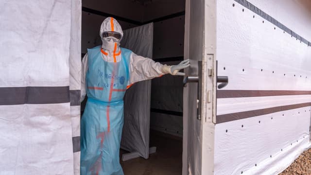 Un soignant en combinaison de protection intégrale, capuche, lunettes et gants, ouvre la porte d'une unité d'isolement Ebola aux parois de bâche blanche ; son nom, « Dr Nelson », et l'heure « 1.30 PM » sont inscrits au marqueur sur son tablier bleu.