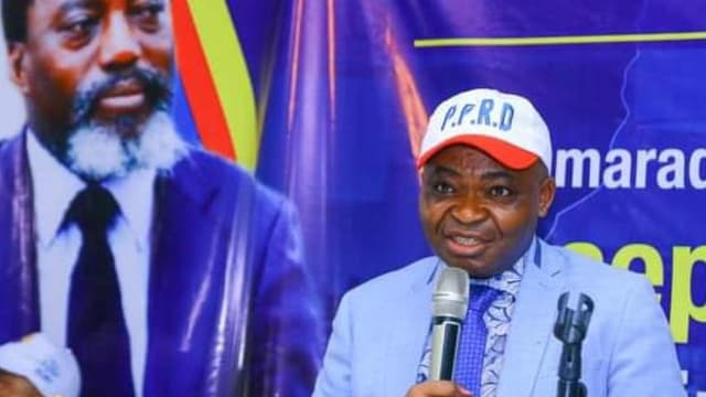 Ferdinand Kambere, secrétaire permanent adjoint du PPRD, parle au micro derrière un pupitre, coiffé d'une casquette blanche siglée P.P.R.D, devant une affiche à l'effigie de Joseph Kabila et une banderole du parti