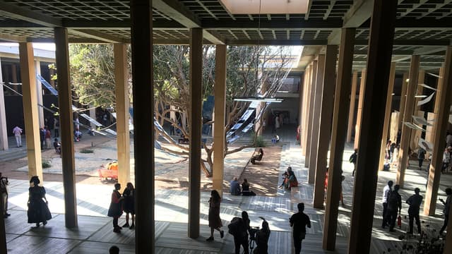 Vue plongeante sur la cour intérieure du site principal de la Biennale de Dakar : un vaste bâtiment ouvert à hautes colonnes, où des visiteurs circulent entre les installations, sous des arbres et des toiles suspendues.