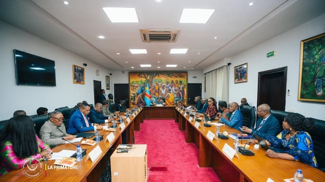 Vue d'une salle de réunion de la Primature à Kinshasa : membres du gouvernement, magistrats et responsables d'institutions assis de part et d'autre d'une longue table en U, face à la table de présidence placée devant une fresque murale aux couleurs du drapeau congolais.
