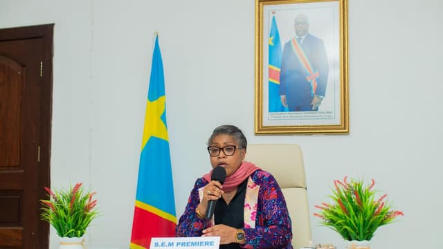 La Première ministre Judith Suminwa Tuluka s'exprime au micro derrière un pupitre « S.E.M Première ministre », devant le drapeau de la RDC et le portrait du président Tshisekedi.