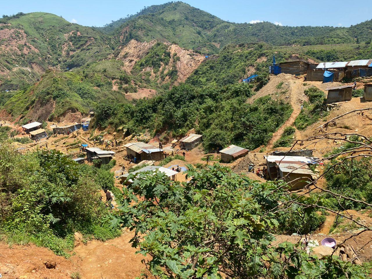 Vue en plongée du site minier artisanal de Lupupu, à Kamituga : des cabanes en planches et en tôle dispersées sur des collines éventrées par des excavations de terre ocre, entourées de végétation verte, avec des creuseurs au travail.