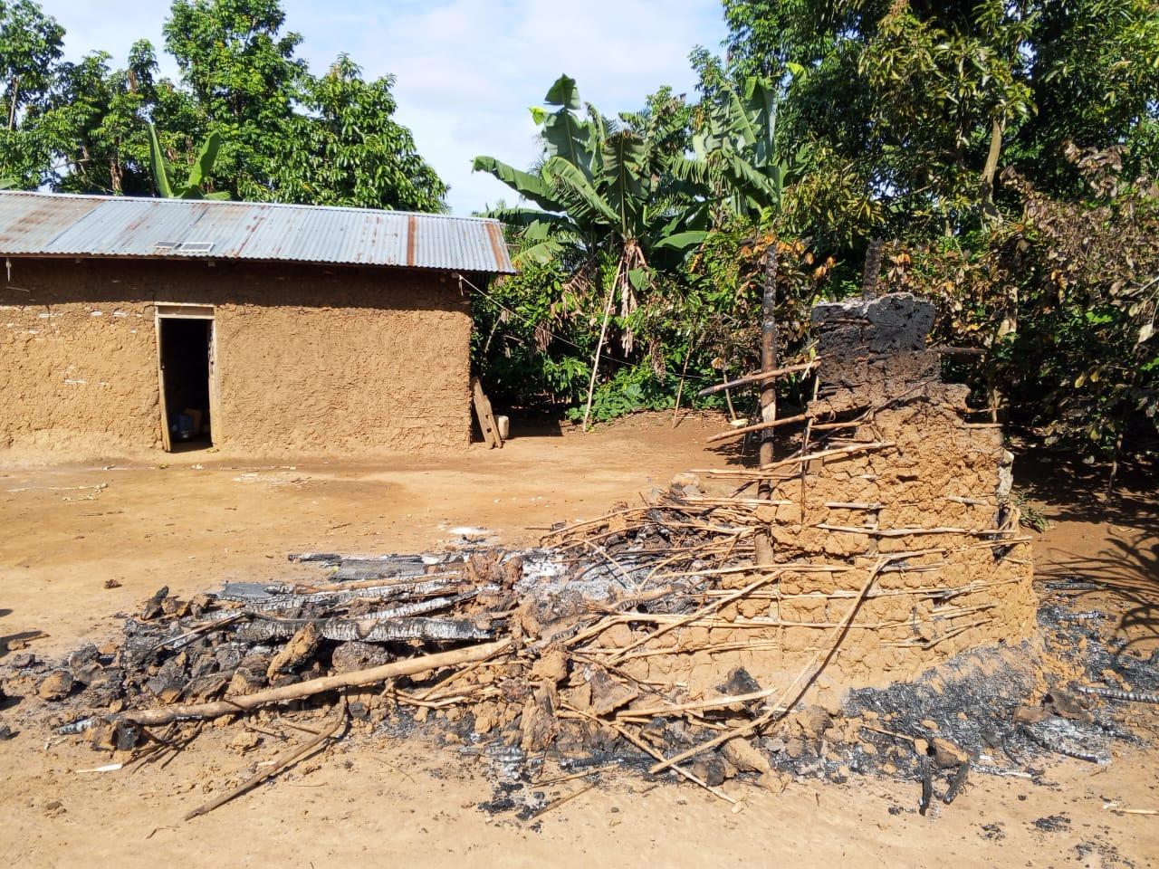 Une maison en briques de terre au toit de tôle et, au premier plan, les décombres calcinés d'une habitation incendiée, dans un village rural entouré de bananiers et de forêt, après une attaque des ADF.