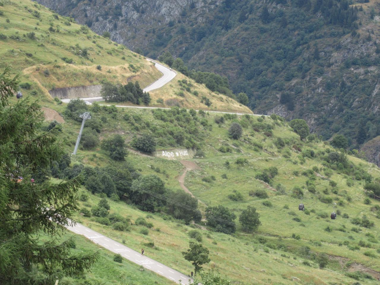 Les lacets en épingle de la montée de l'Alpe d'Huez à flanc de montagne, avec l'inscription « Alpe d'Huez » sur la pente et des cyclistes sur la route lors d'une étape du Tour de France.