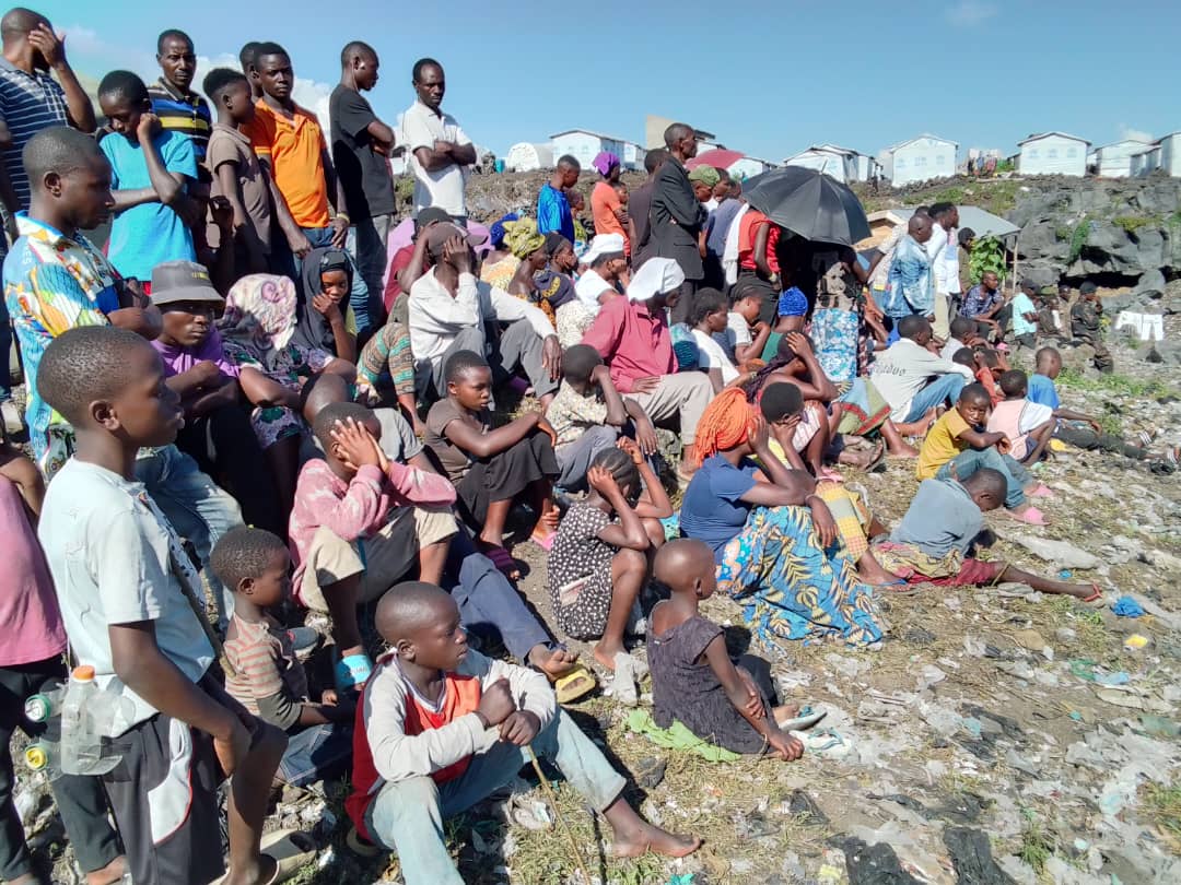 Foule de déplacés congolais, hommes, femmes et enfants, assis à même le sol rocailleux devant des abris de fortune sur un site de déplacés dans l'Est de la RDC.
