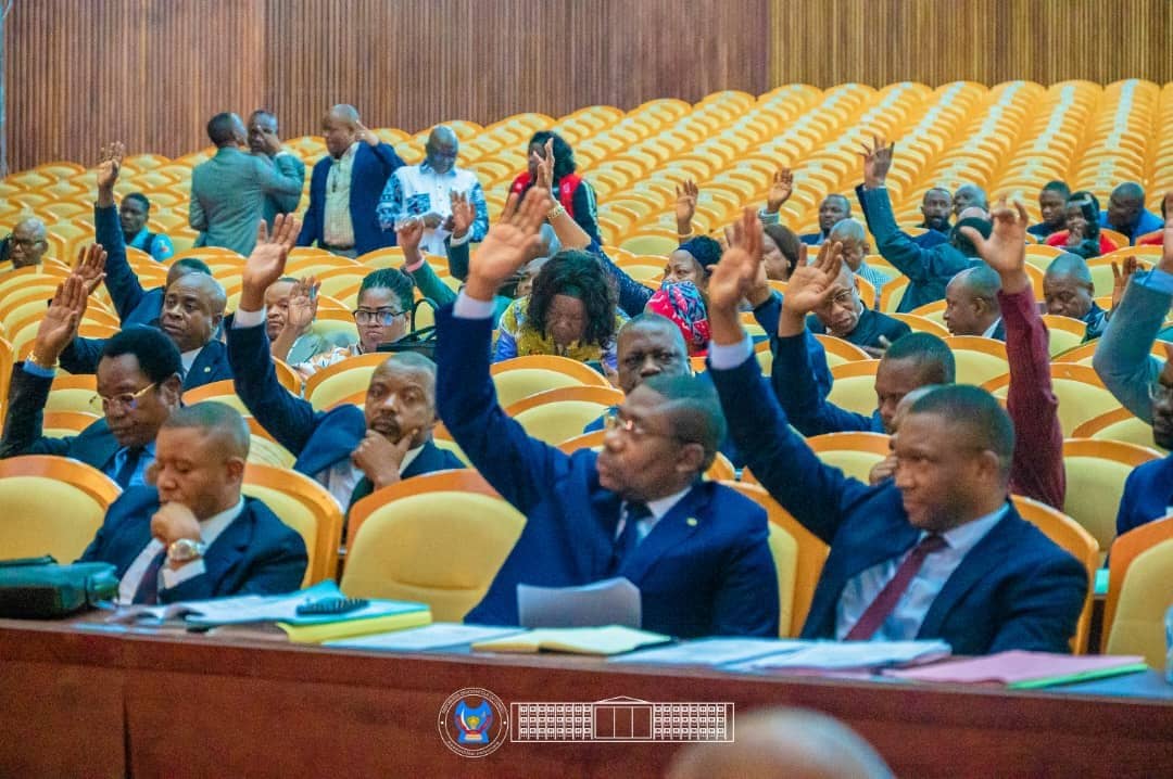 Des députés nationaux, assis dans l'hémicycle du Palais du Peuple à Kinshasa, lèvent la main pour voter lors d'une plénière de l'Assemblée nationale.