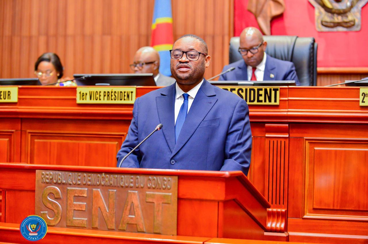 Le ministre congolais des Finances Doudou Fwamba, en costume bleu, s'exprime debout au pupitre marqué « République démocratique du Congo , Sénat », devant les membres du bureau assis à la tribune.