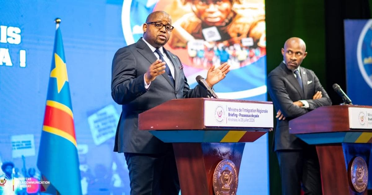 Floribert Anzuluni, ministre de l'Intégration régionale, s'exprime les mains ouvertes derrière un pupitre marqué « Ministre de l'Intégration Régionale, Briefing - Processus de paix, Kinshasa, le 28 juillet 2026 » ; à droite, Patrick Muyaya, bras croisés, à son propre pupitre, devant le drapeau de la RDC.