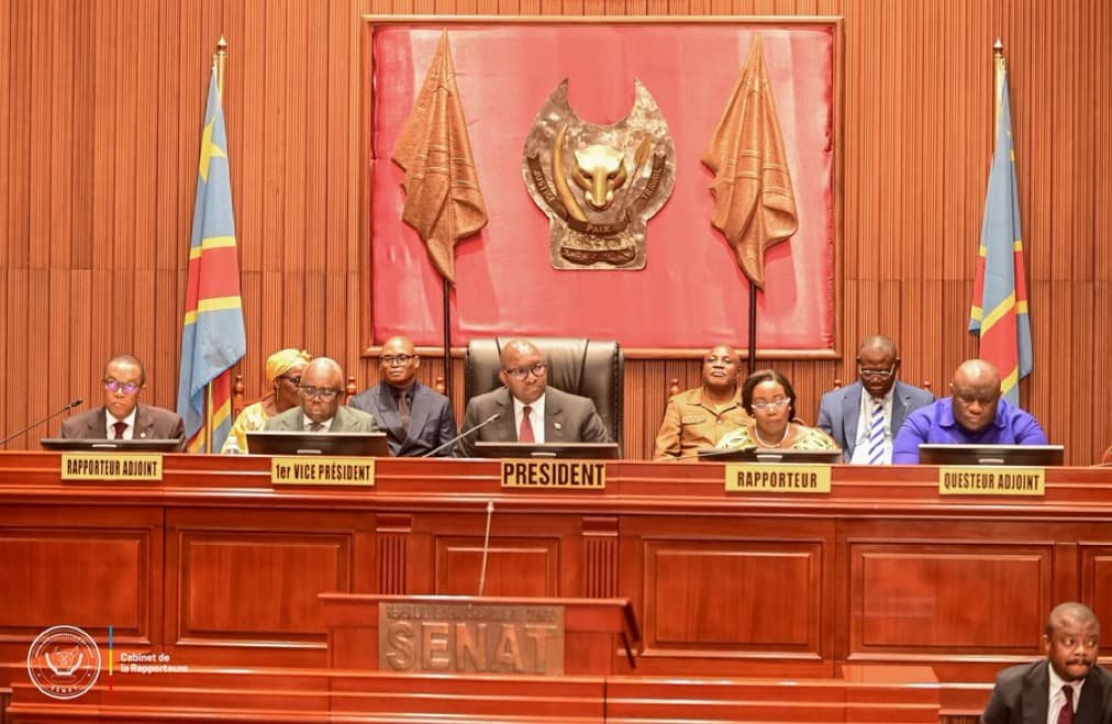 Le présidium du Sénat congolais au Palais du Peuple : Jean-Michel Sama Lukonde, en costume bleu et lunettes, siège au centre sous le pupitre marqué PRÉSIDENT, entouré des vice-présidents, rapporteurs et questeur, devant l'emblème du Sénat et deux drapeaux de la RDC.