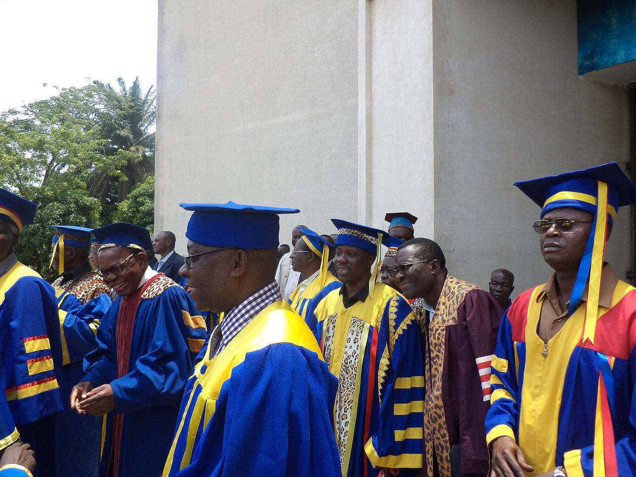 Un groupe de professeurs d'université en toge bleue, jaune et rouge, coiffés de la toque académique, réunis devant un bâtiment de l'Université de Kinshasa.