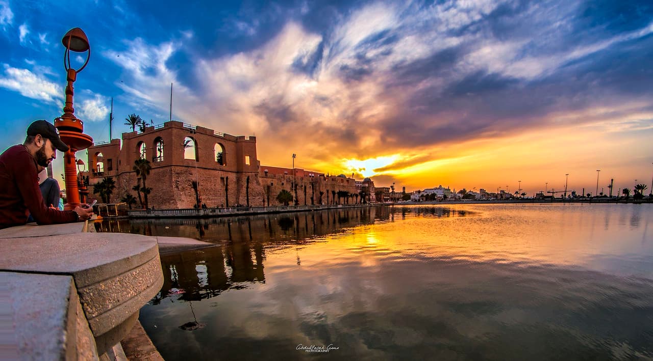 Le château rouge de Tripoli, forteresse de pierre ocre illuminée au coucher du soleil, bordant un plan d'eau qui reflète le ciel orangé ; la ville s'étend à l'arrière-plan.