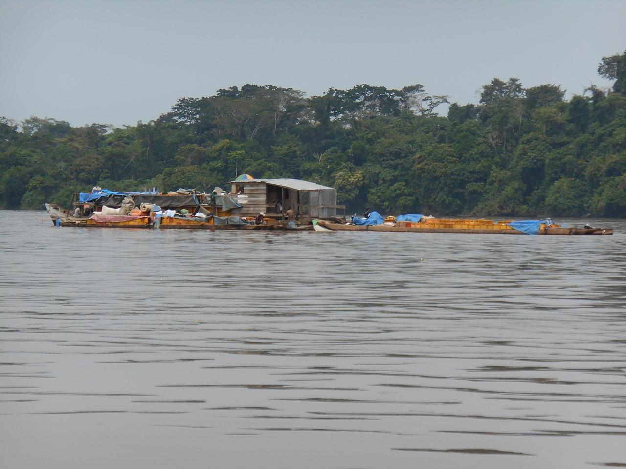 Une longue embarcation en bois, surmontée d'une cabine à toit de tôle et chargée de sacs, de caisses et de bâches, navigue avec des passagers à bord sur un large fleuve bordé de forêt dense.